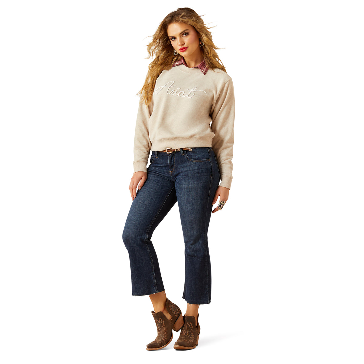 Woman wearing a beige sweater with 'Ariat' branding, blue jeans, and brown boots on a white background.