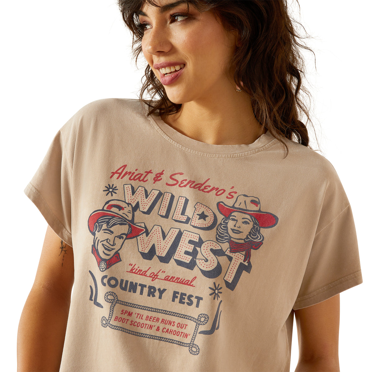 Ariat Women's Sendero x Wild West Country Fest S/S T-Shirt. Woman wearing a beige t-shirt with 'Wild West Country Fest' graphic design.
