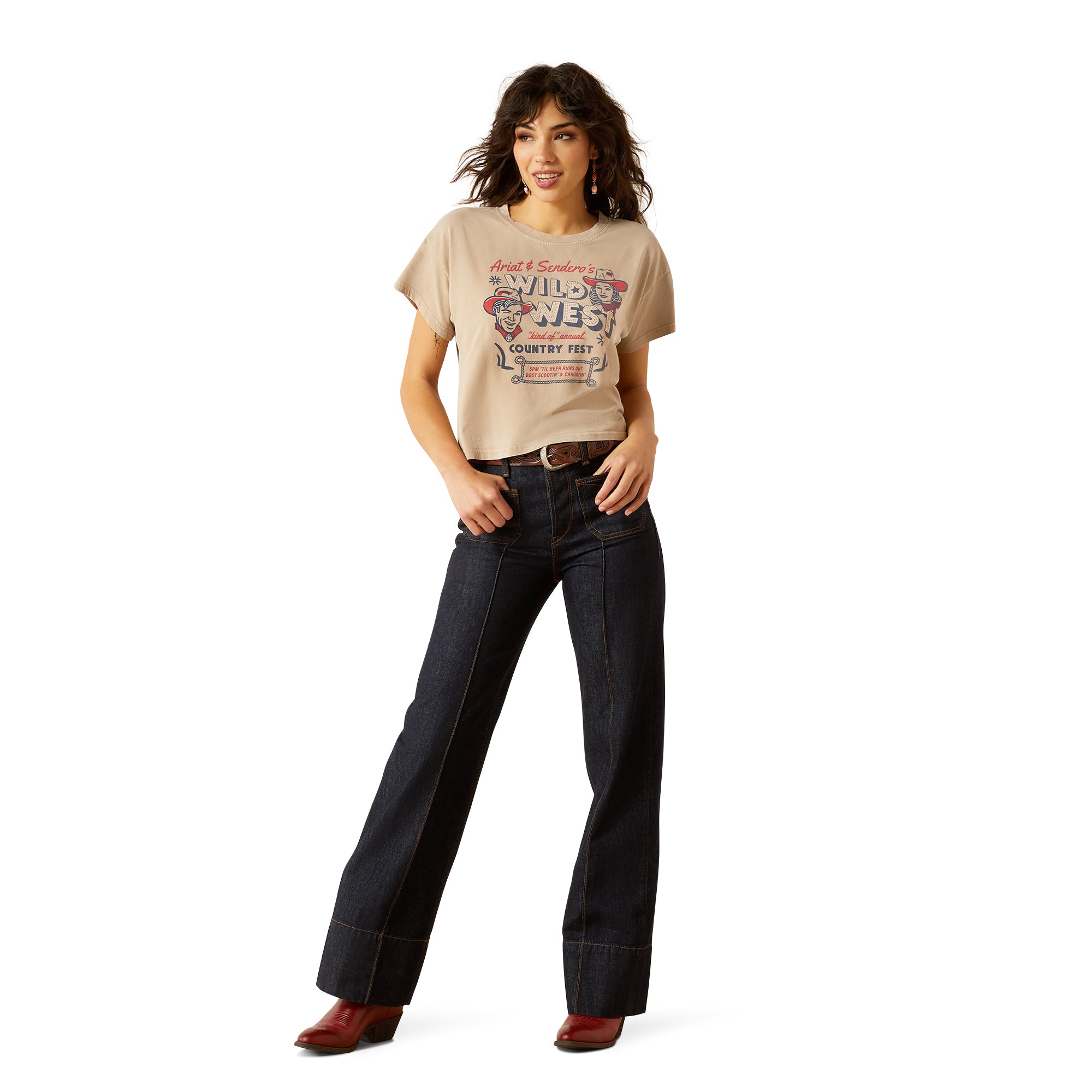 Ariat Women's Sendero x Wild West Country Fest S/S T-Shirt. Woman wearing a beige t-shirt with graphic design and dark jeans on a white background