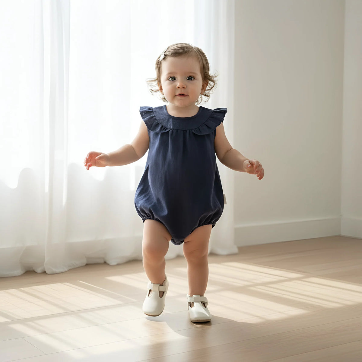 A toddler girl wearing a navy blue ruffle-sleeve romper and white shoes walks barefoot on a wooden floor in front of a bright window with white curtains.