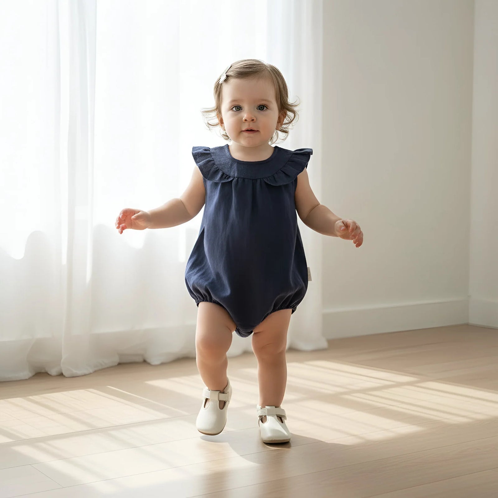 A toddler girl with light brown hair sits cross-legged on a light wood floor in a bright room, wearing a sleeveless navy blue ruffled top and beige baby shoes.
