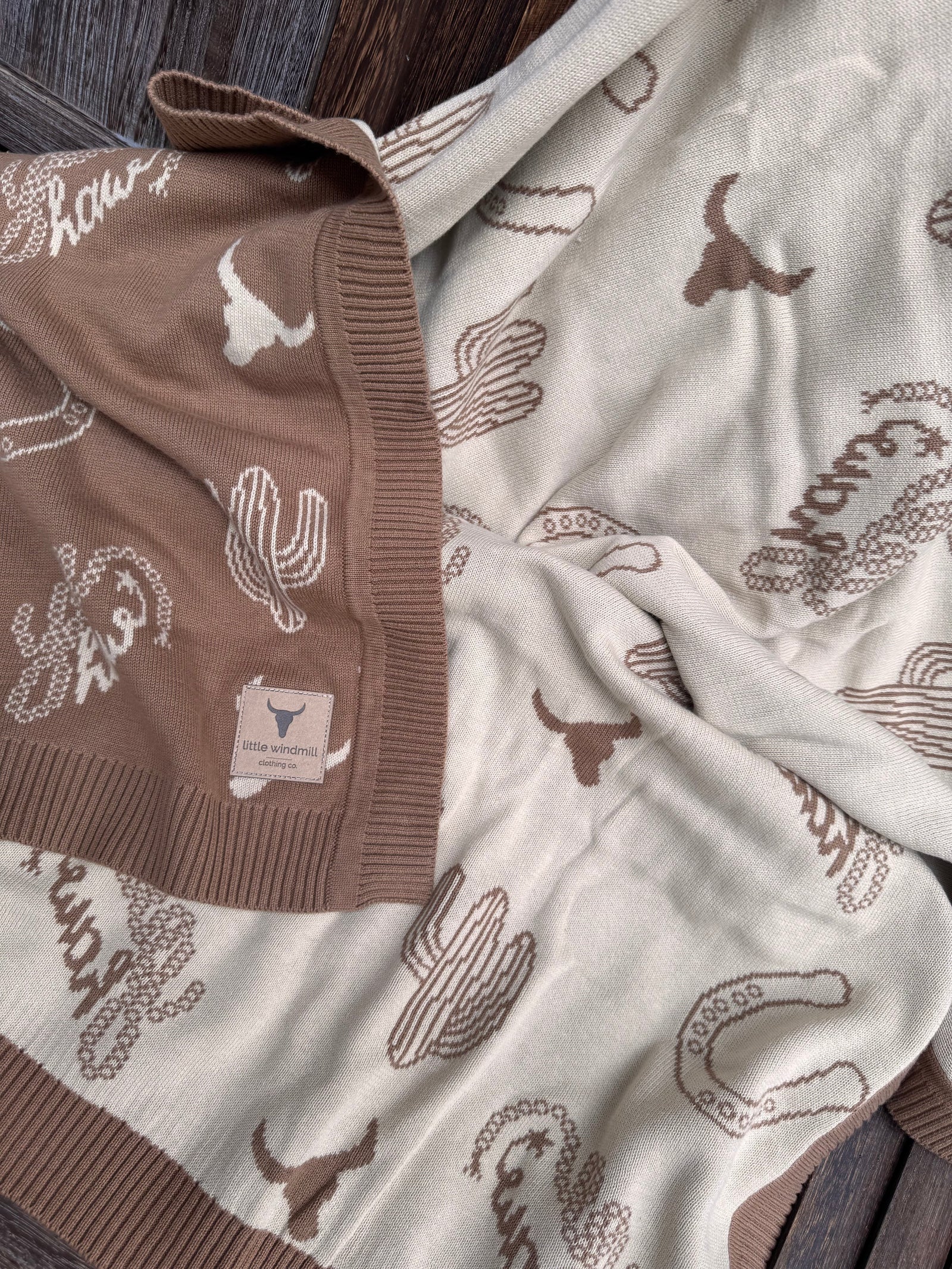 A close-up of a knitted throw blanket with a western theme, featuring brown and cream colors with cactus, longhorn, and horseshoe patterns, laid out on a dark wood surface.