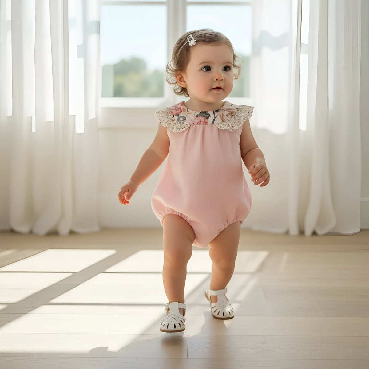 Baby in a pink romper walking in a bright room with white curtains.