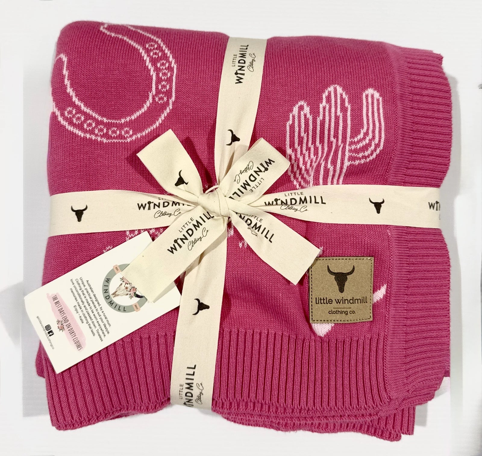 A folded pink knit blanket with white cactus and moon motifs, wrapped in a cream ribbon printed with the Windmill logo, with a brand tag on the corner.