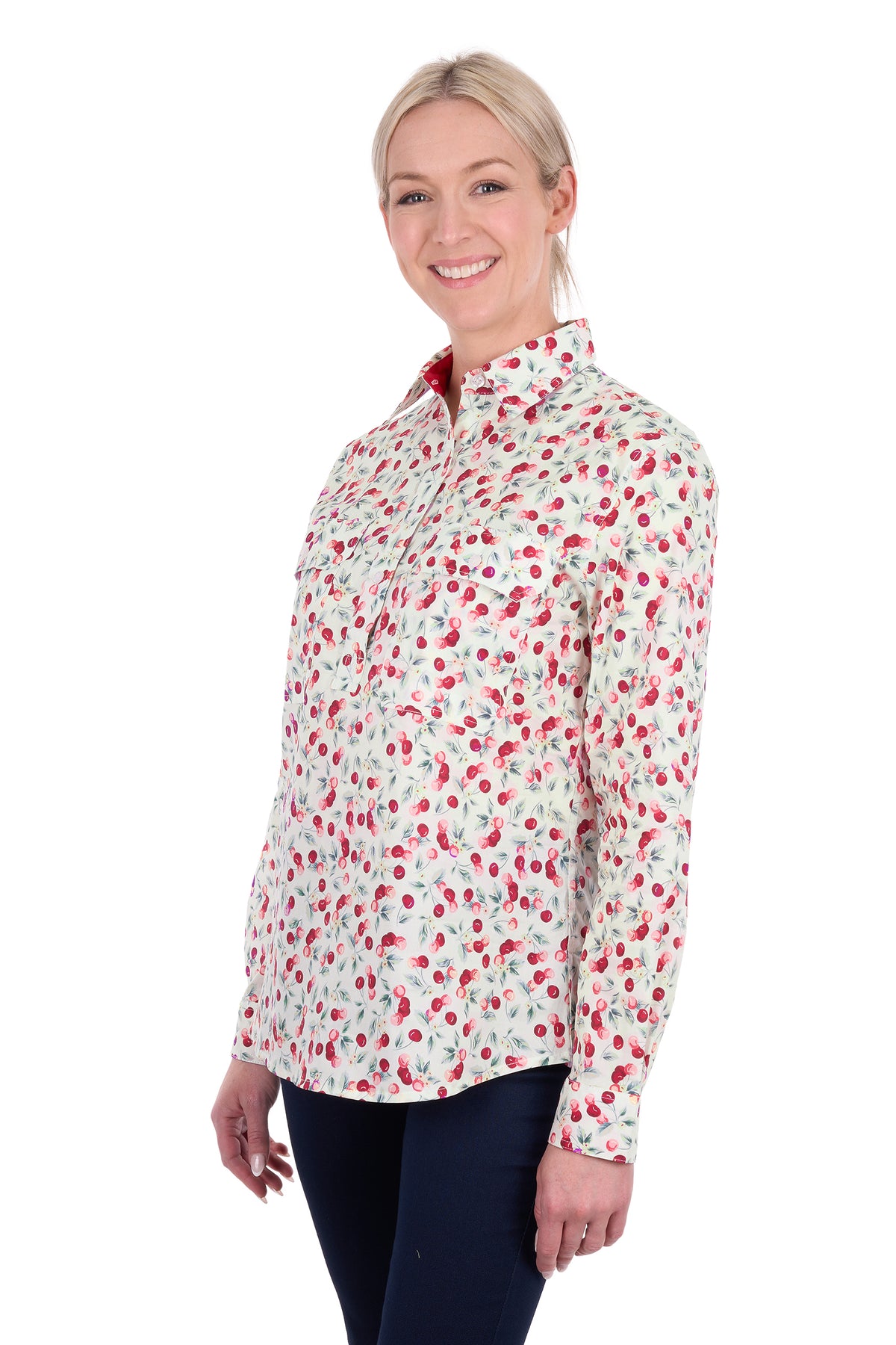 A woman modeling a long-sleeve collared button-up shirt with a red and green cherry print on a white background, paired with dark navy pants, against a plain white studio background.