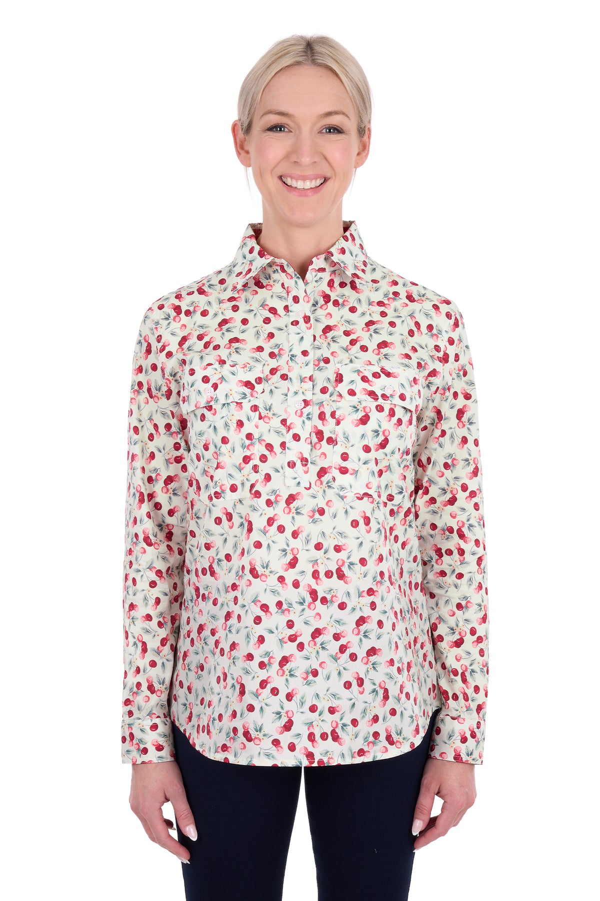 A woman modeling a long-sleeve button-up shirt with a red and green cherry print on a white background, paired with dark pants, against a plain white studio backdrop.