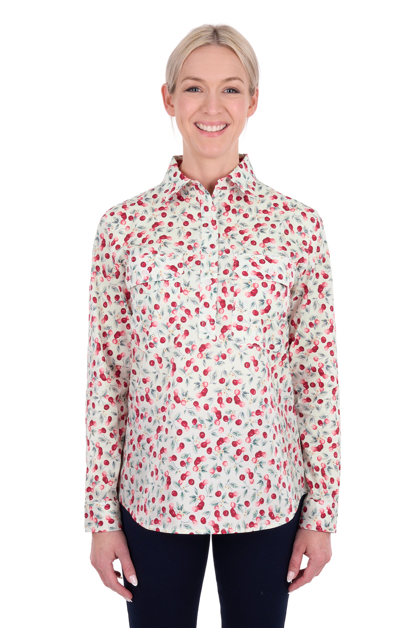 A woman modeling a long-sleeve button-up shirt with a red and green cherry print on a white background, paired with dark pants, against a plain white studio backdrop.