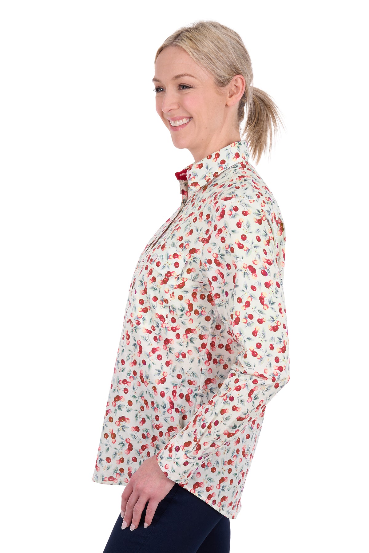 A woman shown in side profile wearing a long-sleeve collared button-up shirt with a red and green strawberry print on a white background, paired with dark navy pants, against a plain white studio background.