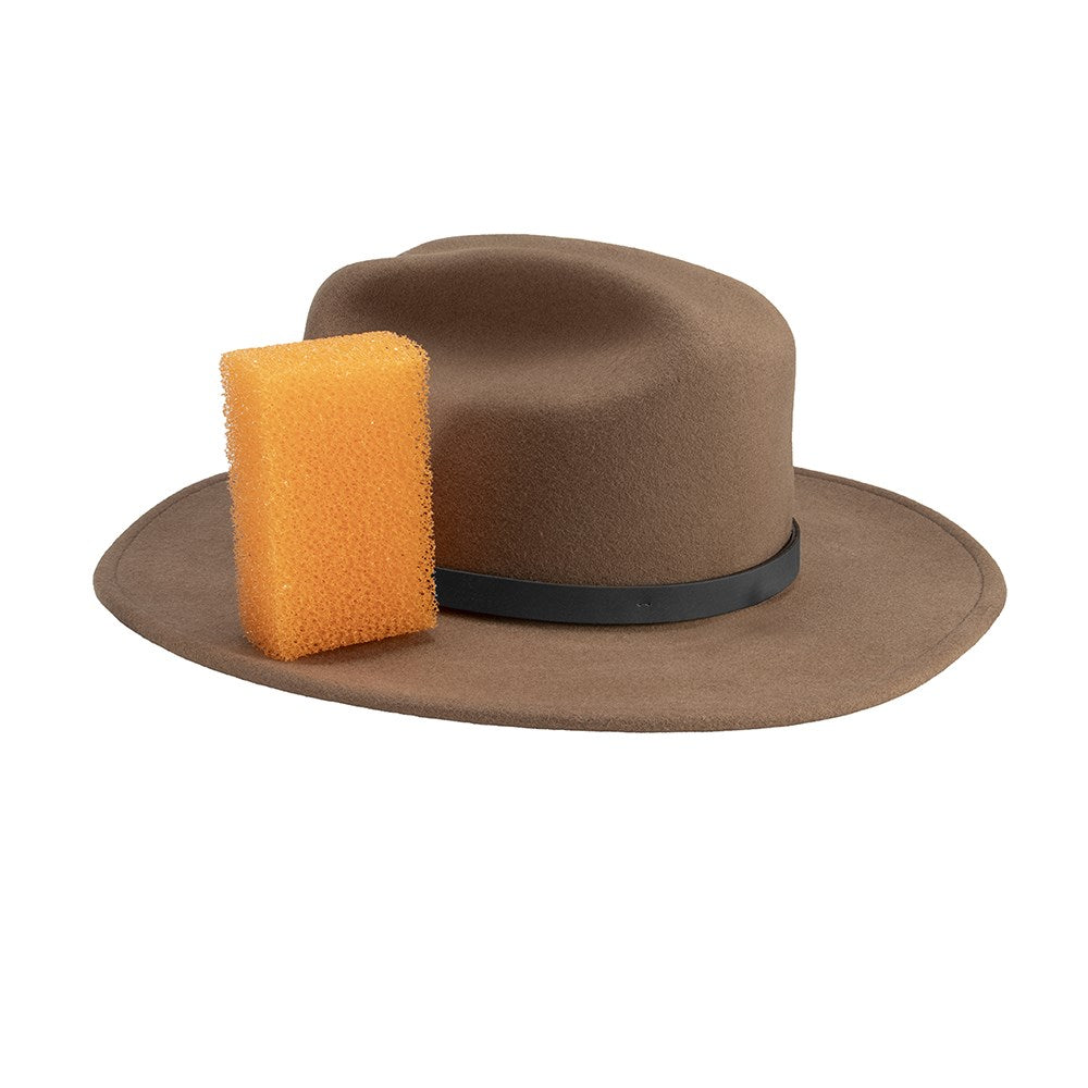 A brown felt fedora hat with a black band and a yellow-orange cleaning sponge attached to the side, shown on a plain white background.