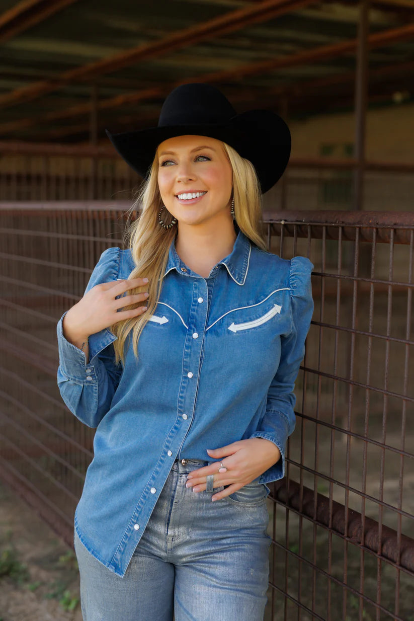 Woman wearing a blue denim shirt with white embroidery and a black cowboy hat in an indoor setting.