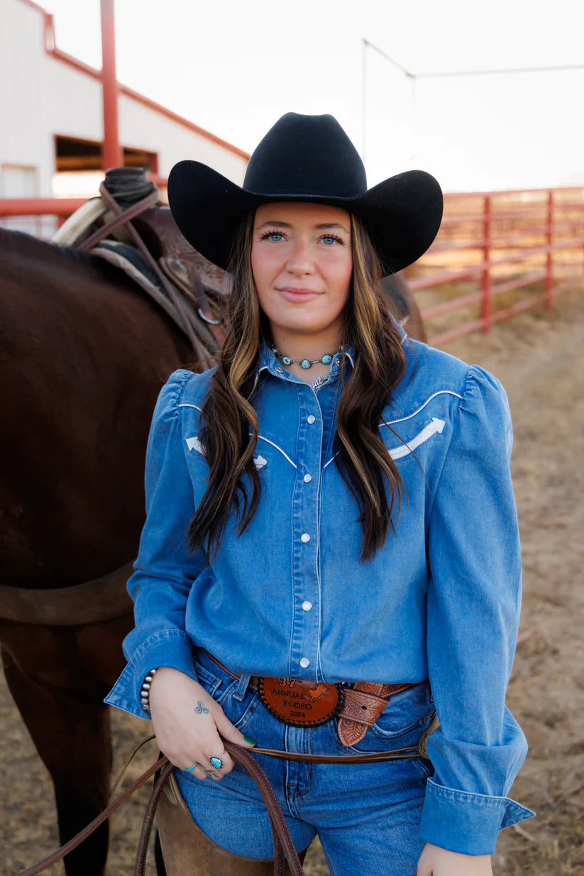 Woman in a blue shirt and cowboy hat standing next to a horse in an outdoor setting