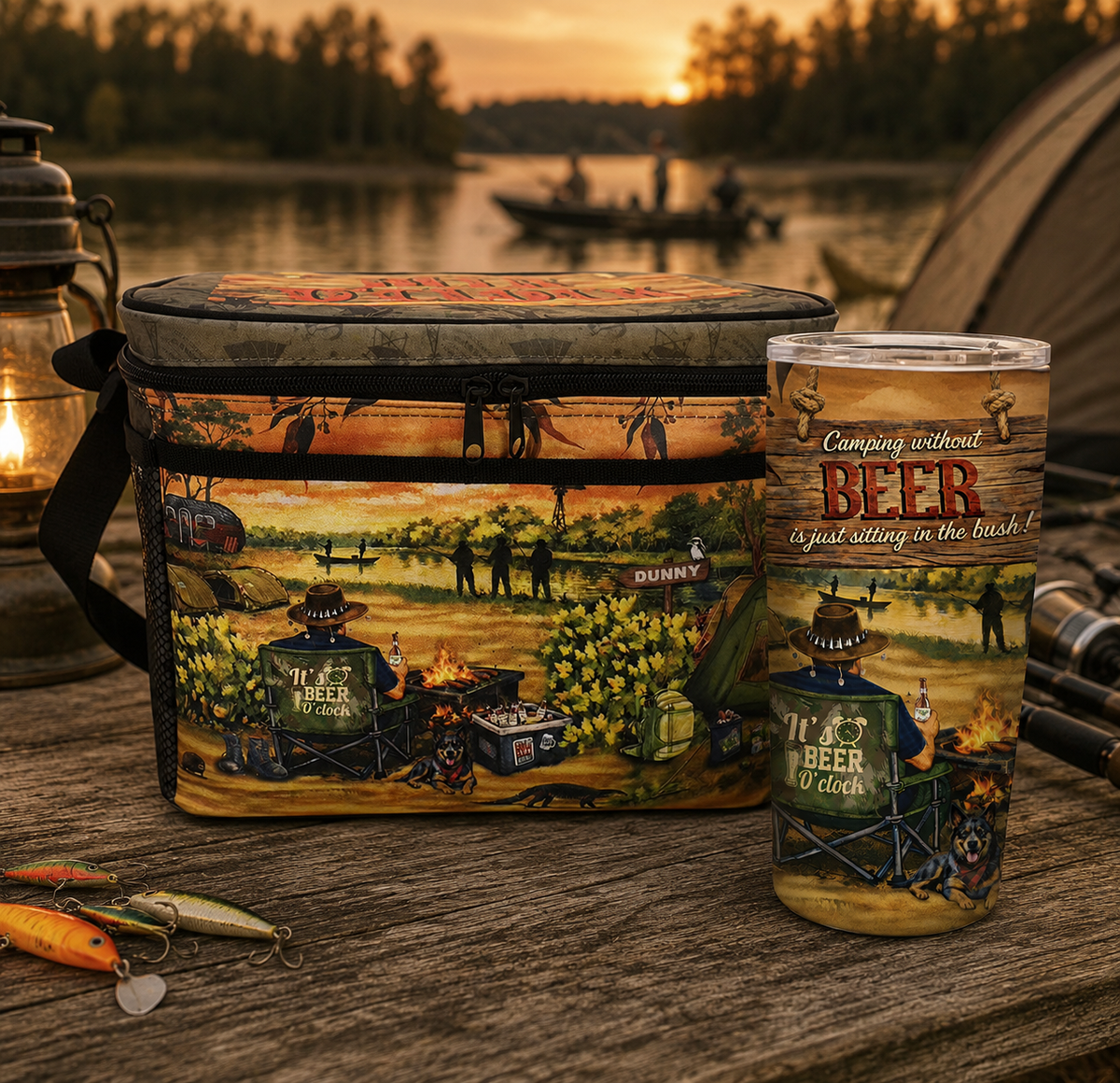 A camping-themed insulated cooler bag and matching tumbler with a rustic forest lake scene printed on both items. The cooler sits on a wooden table outdoors at sunset, with a lantern, fishing lures, tent, and blurred campsite background visible.