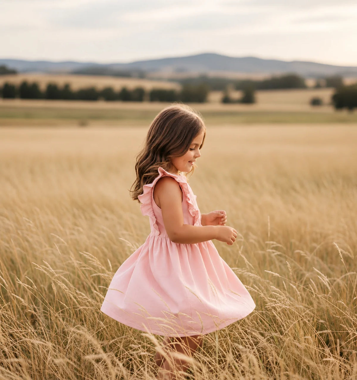 A young girl with long brown hair stands in a golden wheat field wearing a light pink sleeveless dress with ruffled shoulders. The background shows a soft-focus landscape of rolling hills under a warm sky.