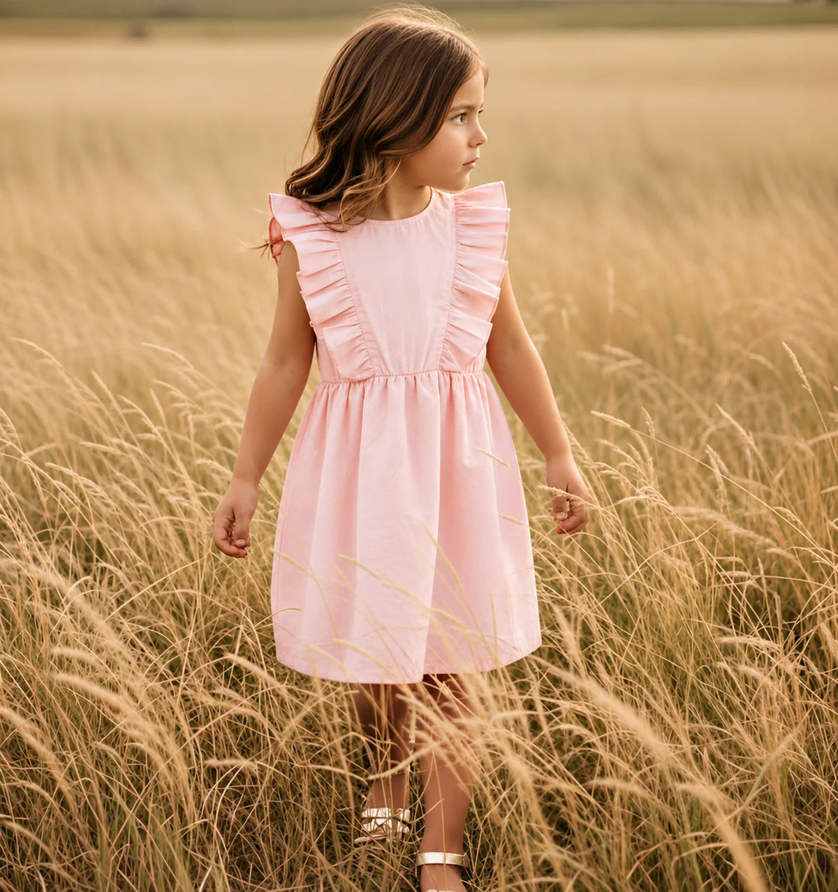 A young girl wearing a light pink sleeveless dress with ruffle details stands in a field of tall golden grass, looking off to the side.