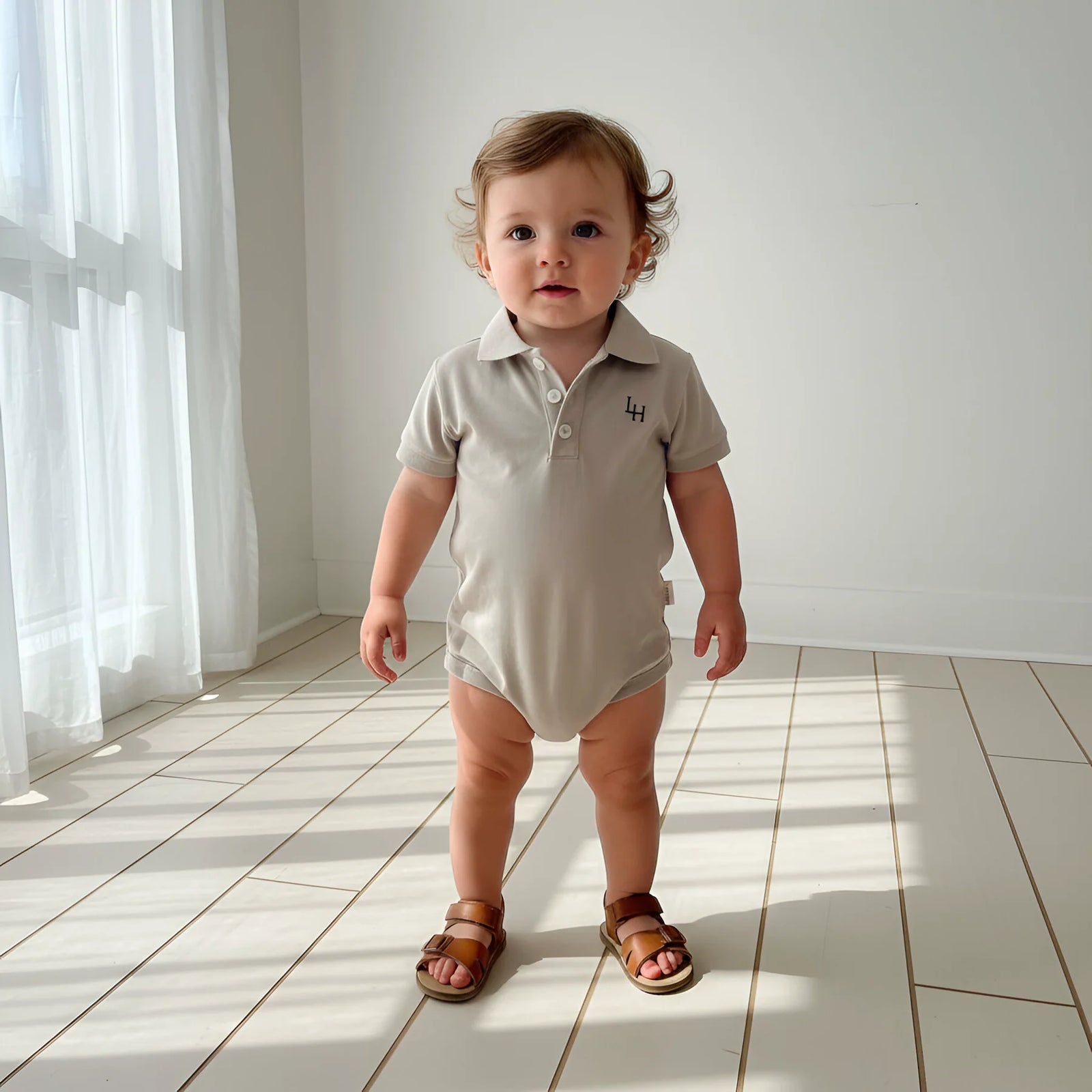 Baby sitting on a wooden floor wearing a beige polo shirt and shorts with brown shoes.