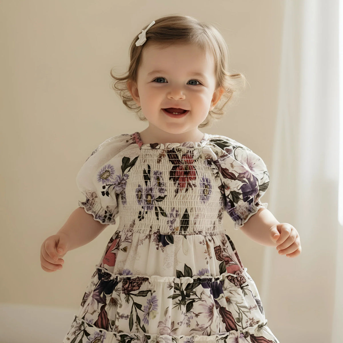A smiling toddler girl wearing a cream-colored smocked dress with a purple, pink, and green floral print, standing against a soft beige background.