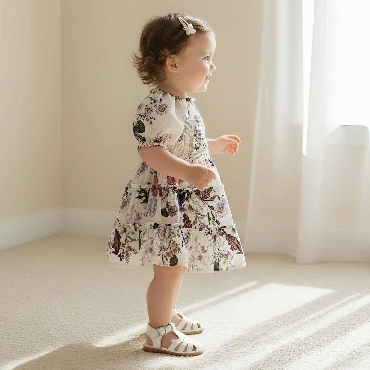 A toddler girl wearing a short-sleeve tiered dress with a floral and smocked bodice, standing in a brightly lit room with beige walls and carpet.