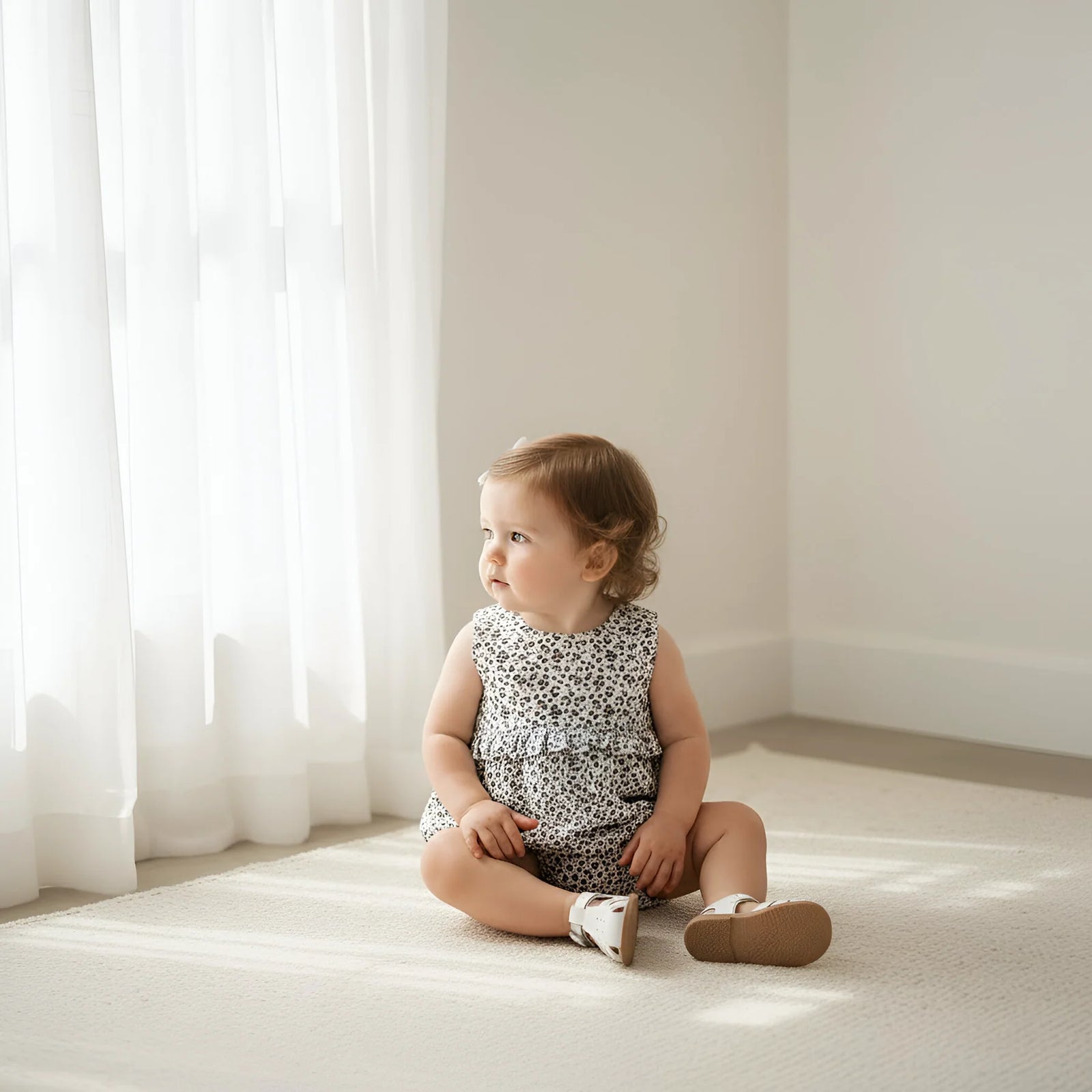 Baby sitting on a light-colored floor in a softly lit room with white curtains.