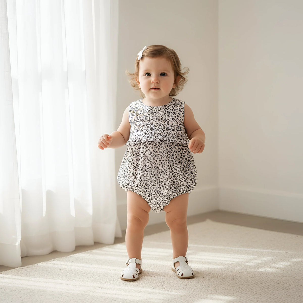 Baby in a floral romper standing in a softly lit room with white curtains.