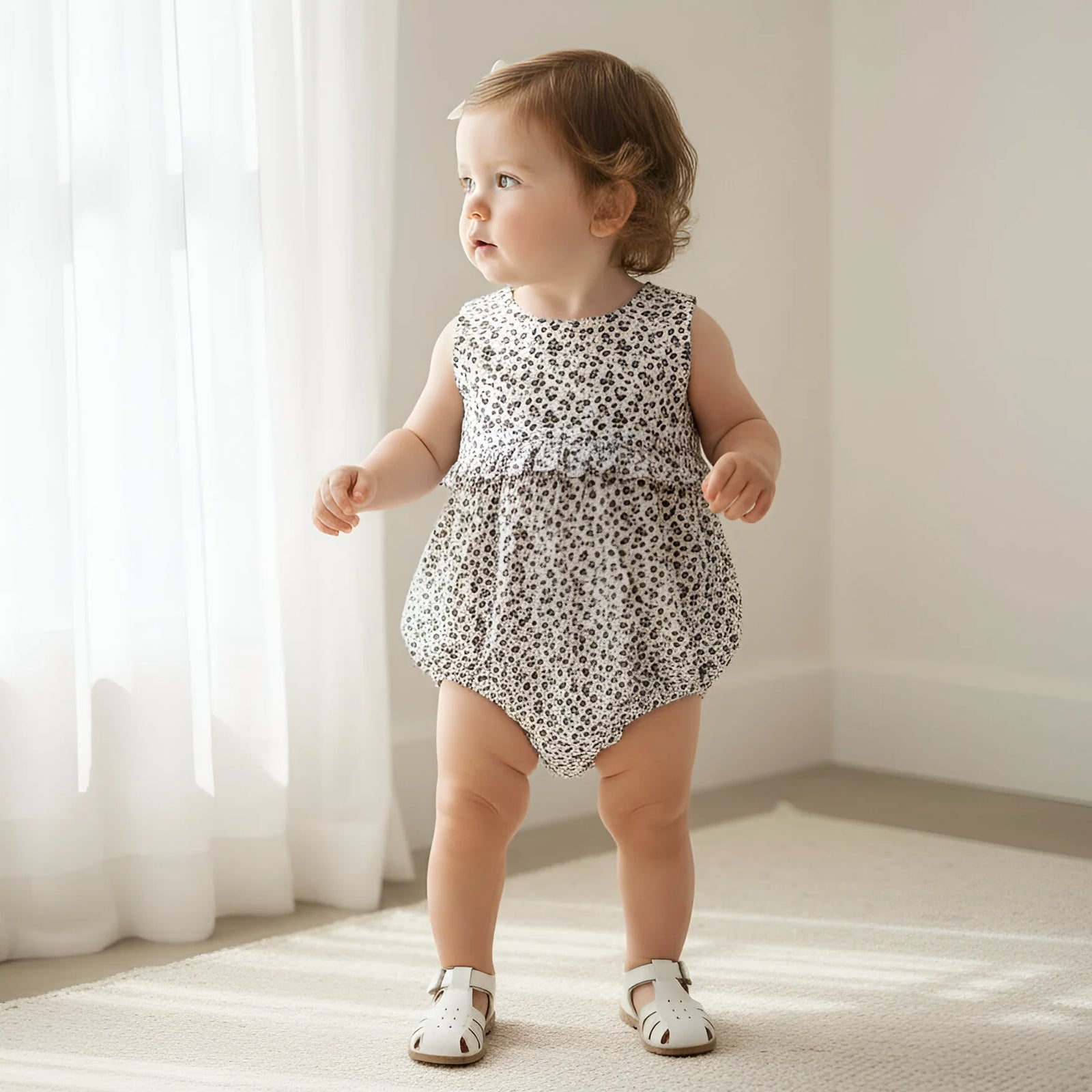 Baby in a patterned romper standing in a softly lit room with white curtains.