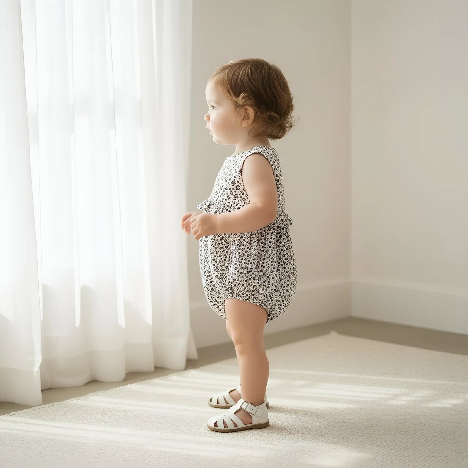 Baby in a patterned romper standing in a softly lit room with white curtains.