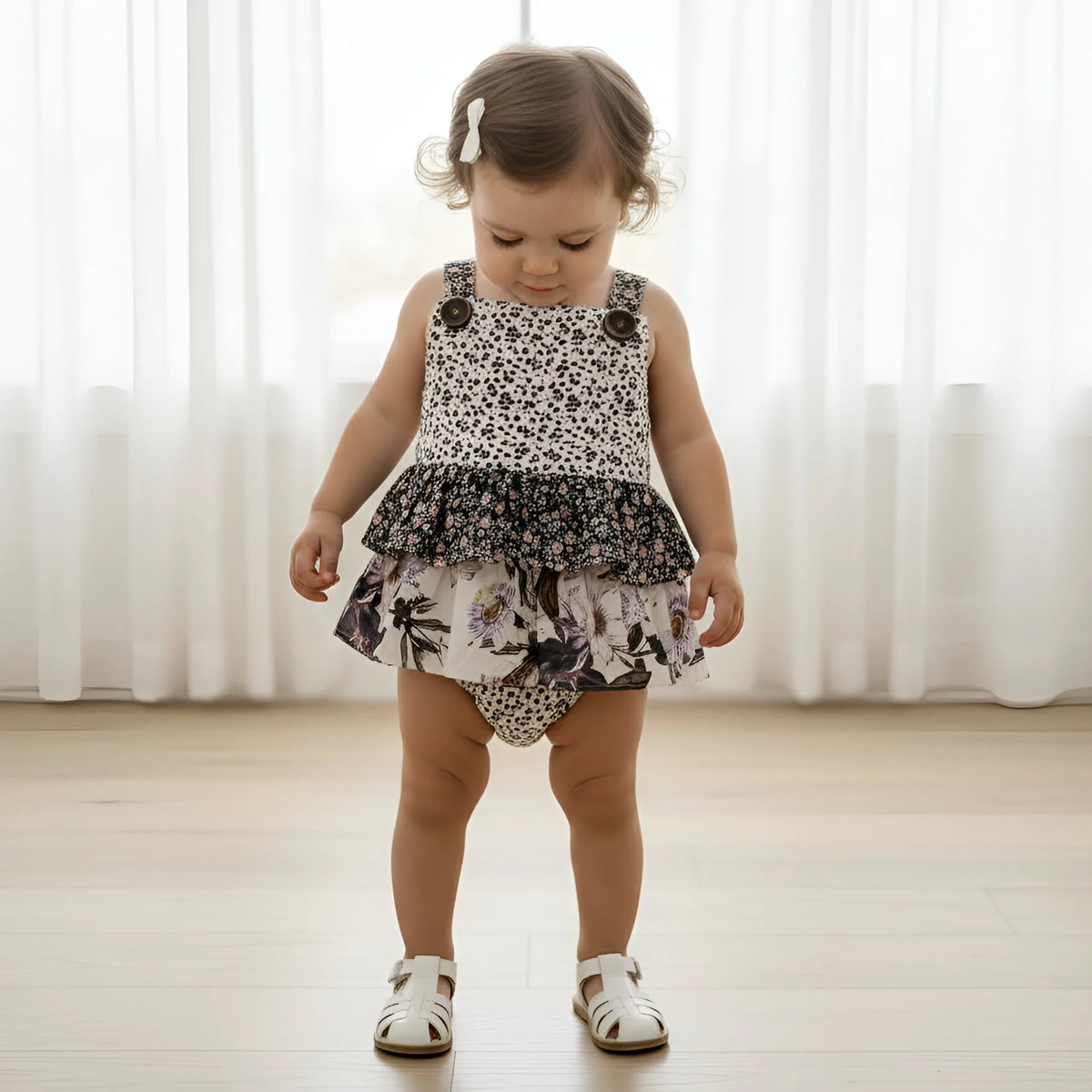 A toddler girl wearing a sleeveless ruffled romper with a black-and-white animal print top and a floral skirt, standing on a light wood floor in front of white curtains.
