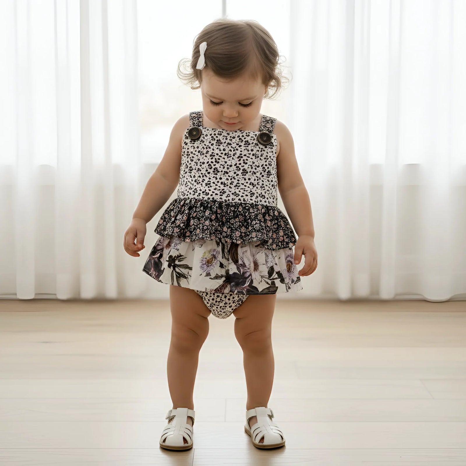 A toddler girl wearing a sleeveless ruffled romper with a black-and-white animal print top and a floral skirt, standing on a light wood floor in front of white curtains.