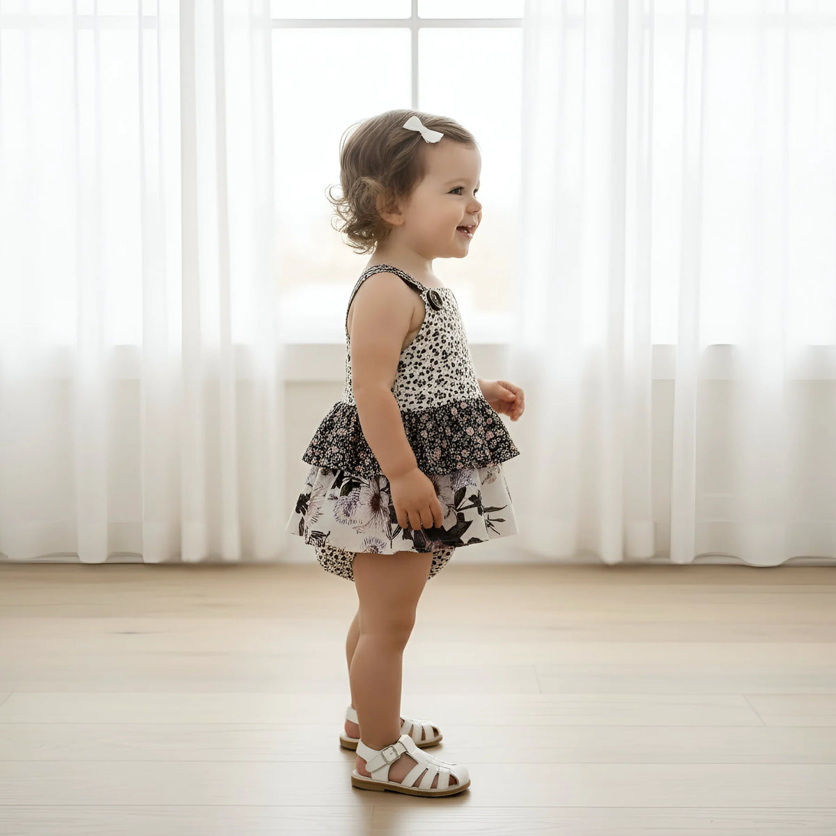 A toddler girl wearing a sleeveless black and white patterned romper dress with a ruffled skirt, standing in profile in a bright room with sheer white curtains and light wood floors.