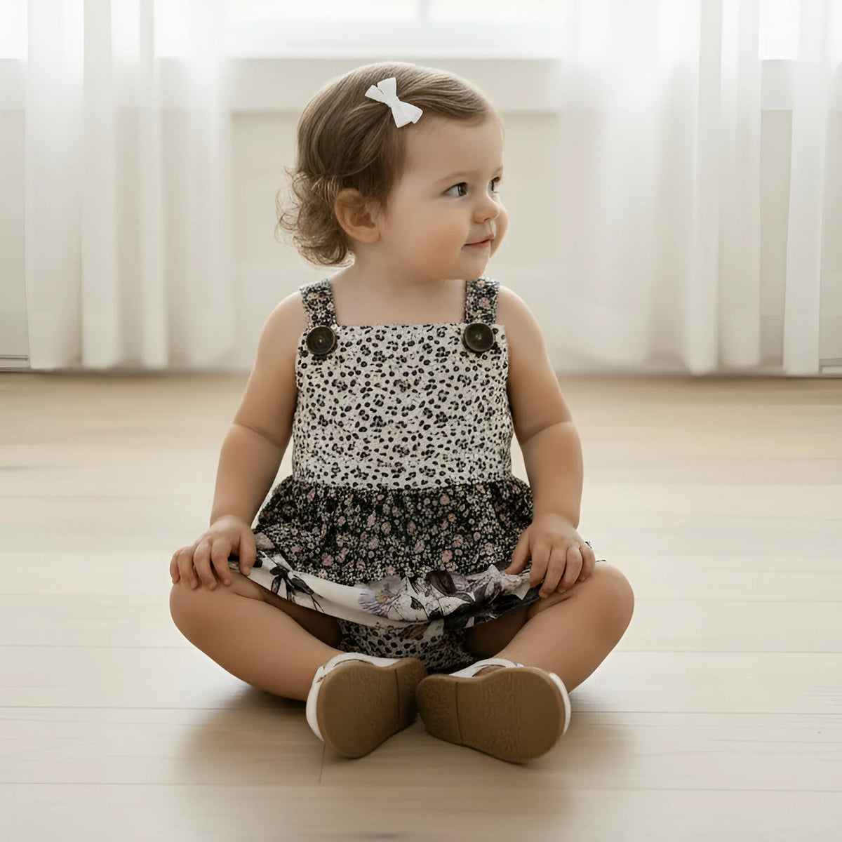 A toddler girl sits cross-legged on a light wooden floor in a bright room with white curtains. She is wearing a sleeveless romper with a black and white animal print pattern, brown shoulder buttons, and a ruffled black skirt.