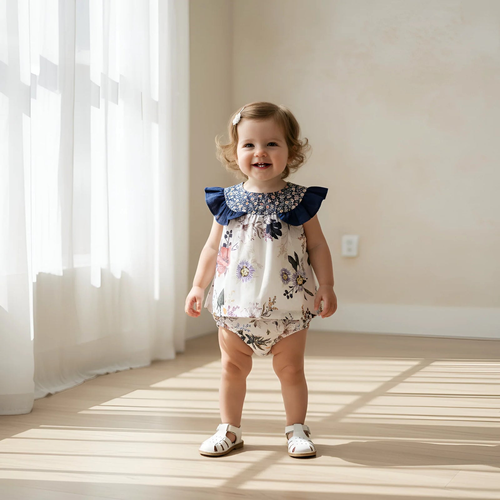 A toddler stands indoors near a window wearing a floral romper with dark blue ruffled shoulder details and white sandals.