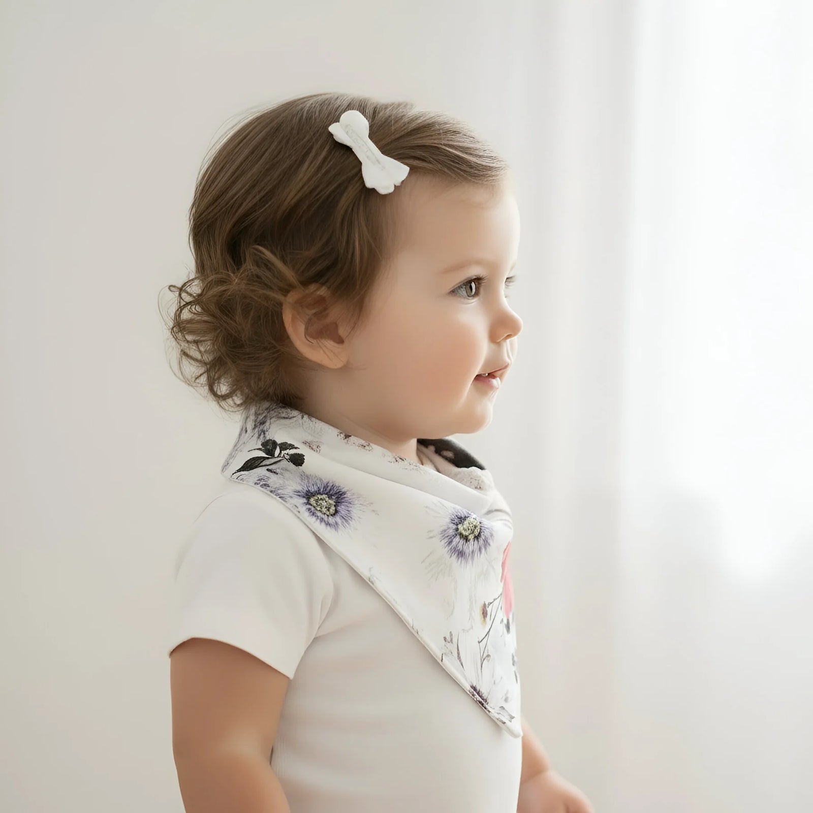 A smiling toddler wearing a white short-sleeve bodysuit and a white triangular bandana bib with a colorful floral print, photographed against a soft white curtain background.