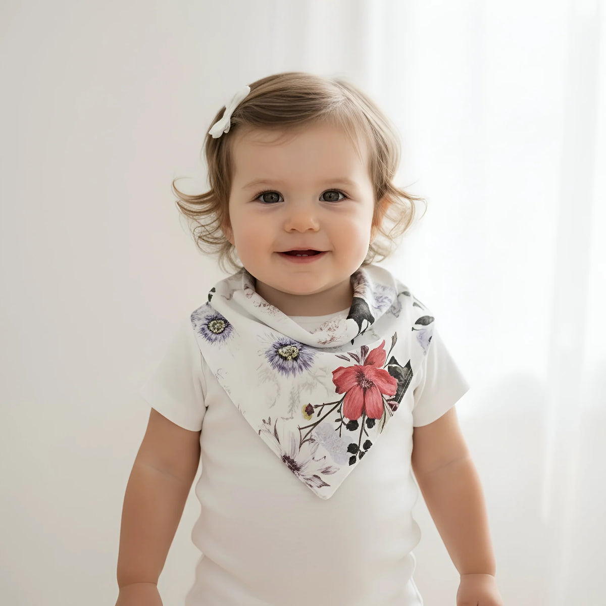A smiling toddler wearing a white short-sleeve bodysuit and a white triangular bandana bib with a colorful floral print, photographed against a soft white curtain background.