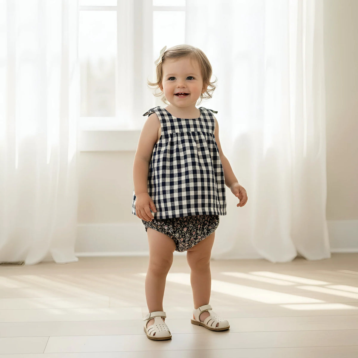 Child wearing a black and white checkered top and floral shorts standing in a bright room with white curtains.
