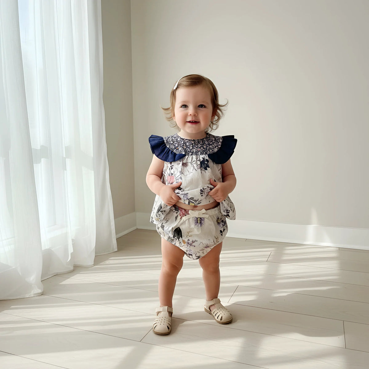 A toddler girl stands in a bright room with sunlight and window curtains, wearing a floral print romper with a navy blue ruffled collar and beige sandals.