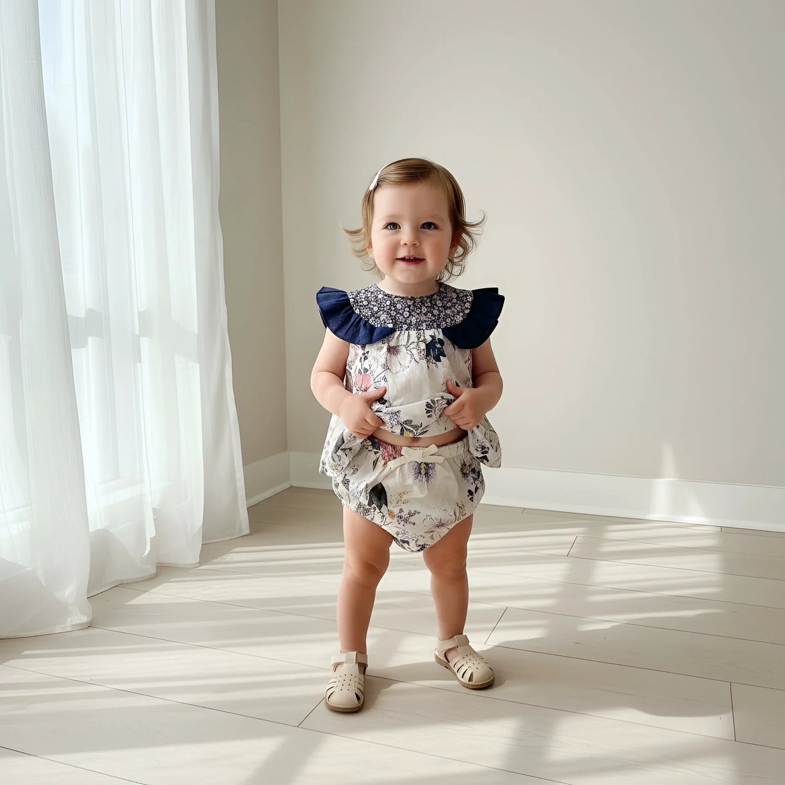 A toddler girl stands in a bright room with sunlight and window curtains, wearing a floral print romper with a navy blue ruffled collar and beige sandals.