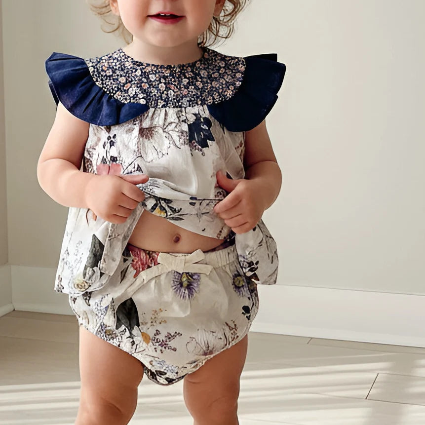 A toddler wearing a two-piece floral romper set with a navy blue ruffled collar and matching floral bloomers, standing in a brightly lit indoor room.