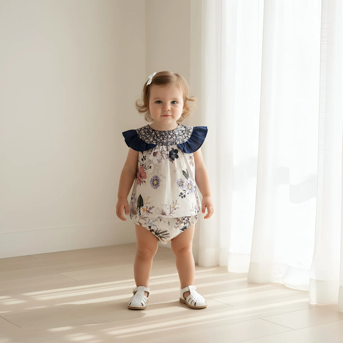 A toddler girl stands in a bright room with white curtains and sunlight on the floor, wearing a sleeveless floral romper with navy ruffled shoulders and white sandals.