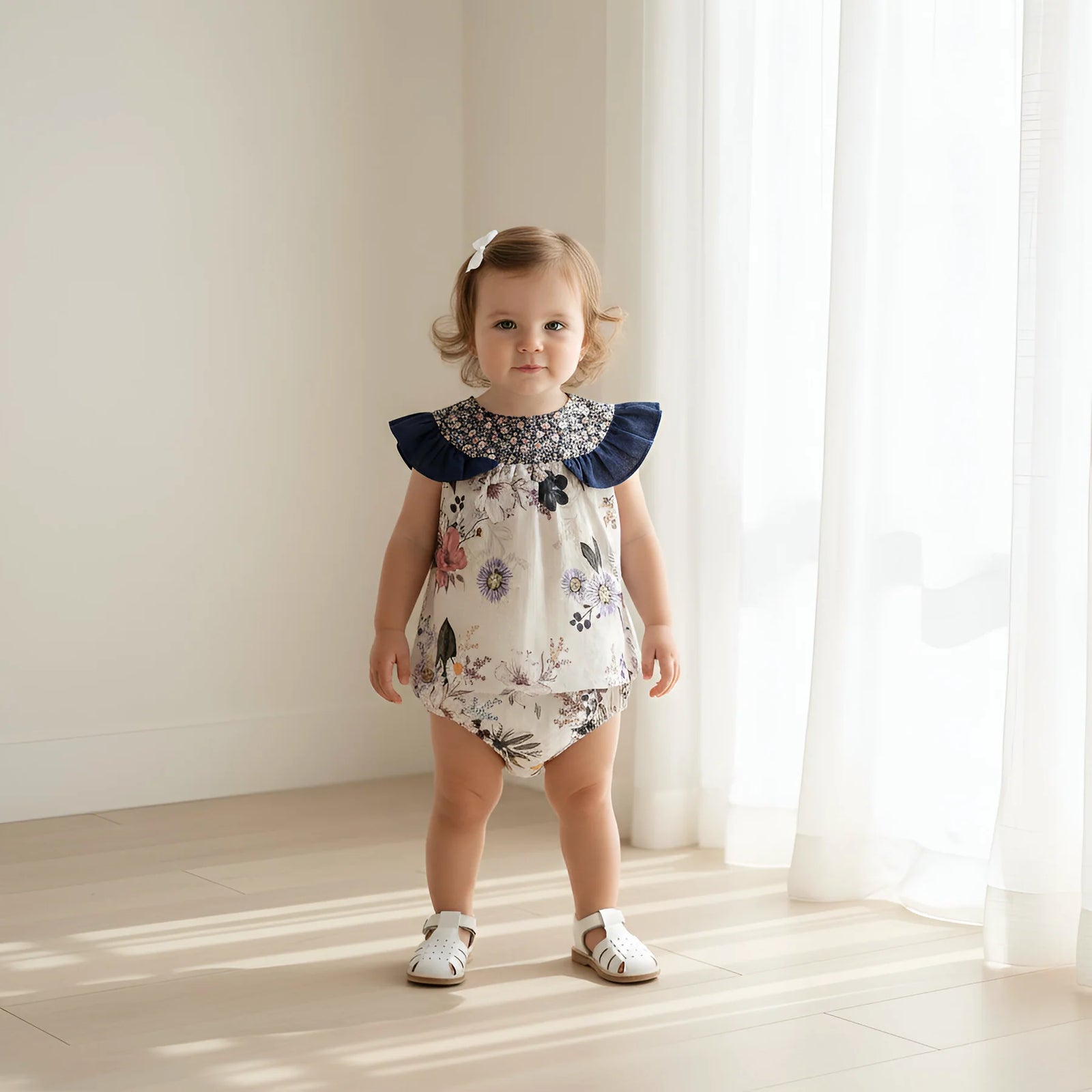 A toddler girl stands in a bright room with white curtains and sunlight on the floor, wearing a sleeveless floral romper with navy ruffled shoulders and white sandals.