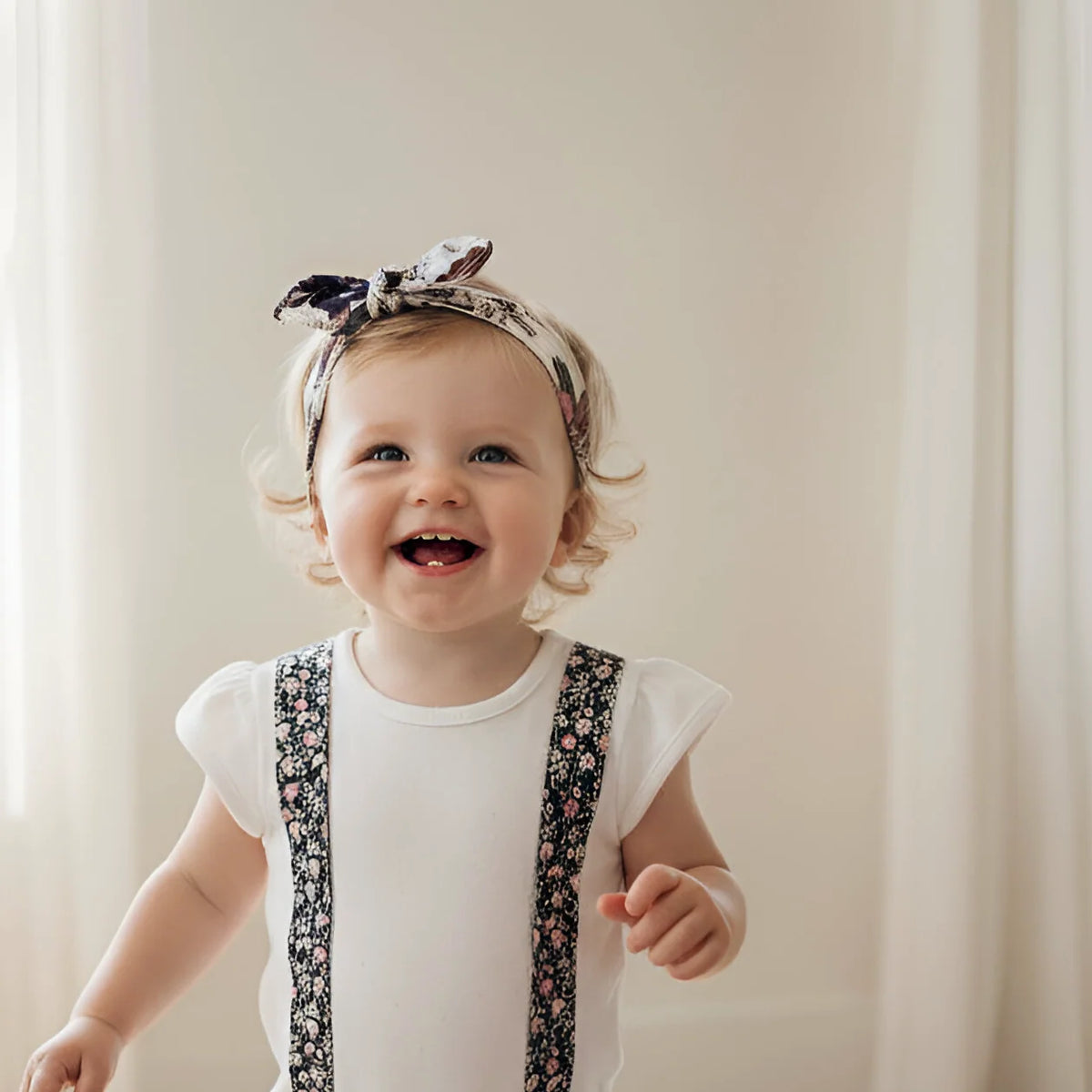 A smiling toddler wearing a floral-patterned headband with a bow and matching floral suspenders over a white short-sleeve top, standing in front of a soft cream curtain background.
