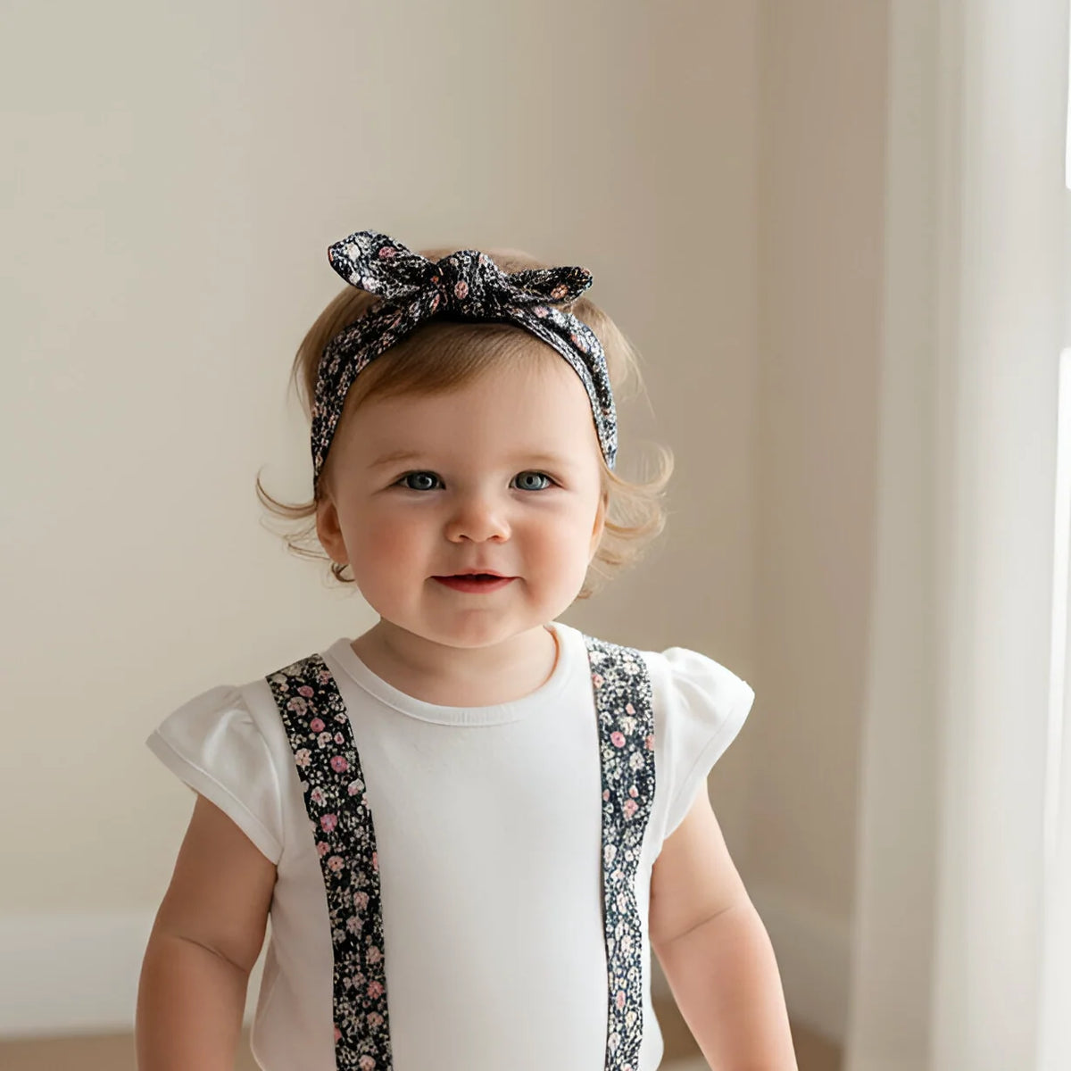 Baby wearing a white onesie with floral details and a headband, standing in a softly lit room.