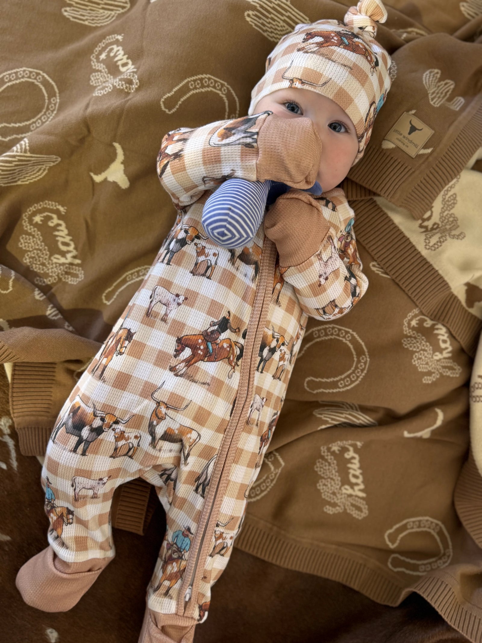 A baby wearing a brown and beige plaid zip-up onesie with a western horse print, matching hat, and blue striped teething toy, lying on a brown blanket with cowboy-themed prints.