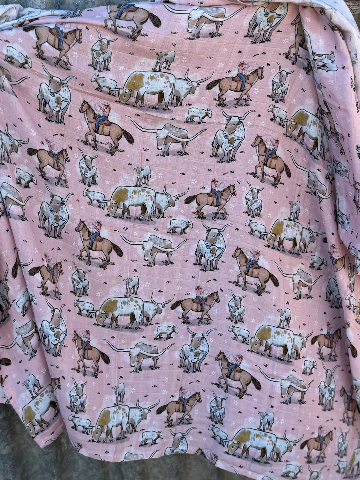A pink blanket or throw with an all-over western-themed print of cattle, horses, and cowboys, draped to show its soft texture and pattern.