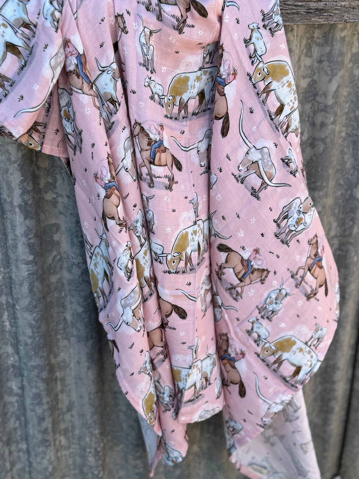 A light pink fabric garment or wrap with an all-over cowboy and cattle print, draped against a weathered gray wooden fence.