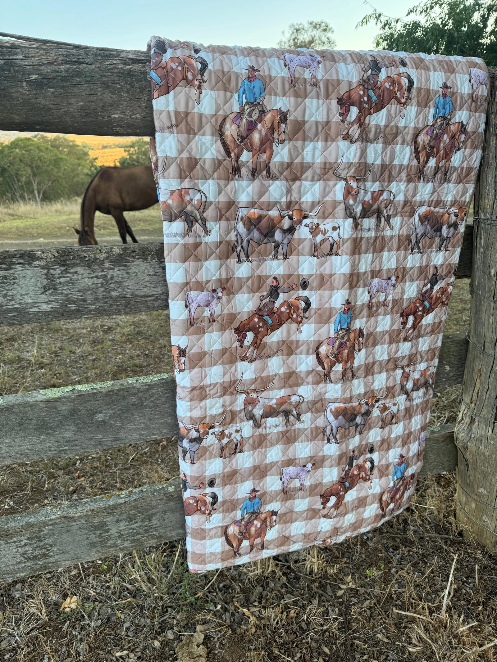 A quilted blanket with a brown and white checkered pattern and cowboy-on-horse motifs, draped over a rustic wooden fence in an outdoor ranch setting with a horse visible in the background.