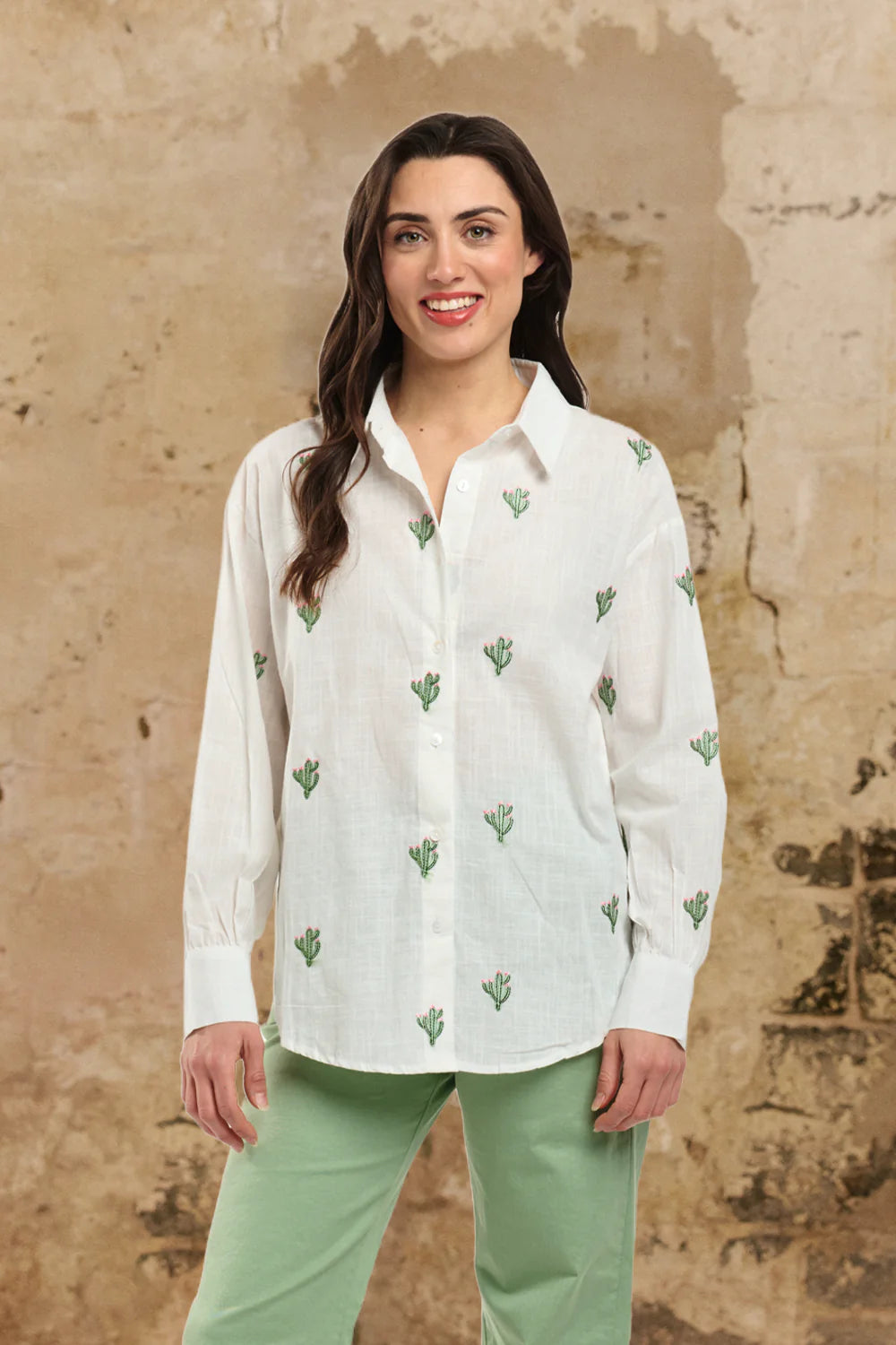 Woman wearing a white blouse with green patterns and green pants against a beige wall.