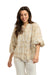 Italian Star Women's Freida Check Shirt. Woman wearing a patterned blouse and white pants standing against a stone wall.