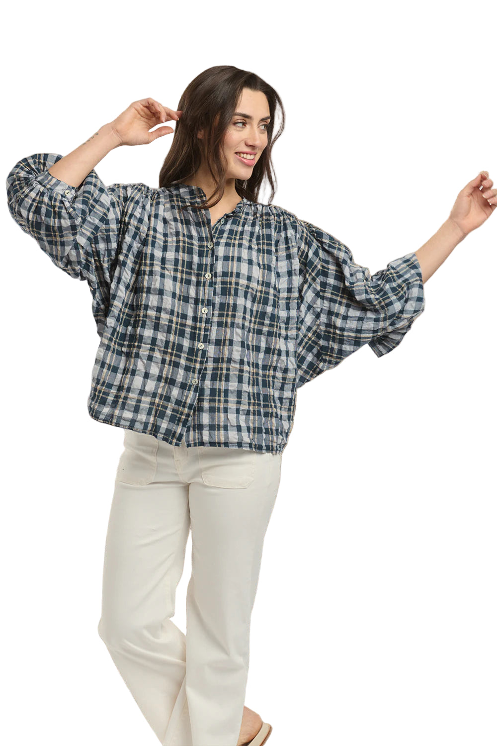 Italian Star Women's Freida Check Shirt. Woman wearing a blue plaid shirt and white pants standing against a stone wall.