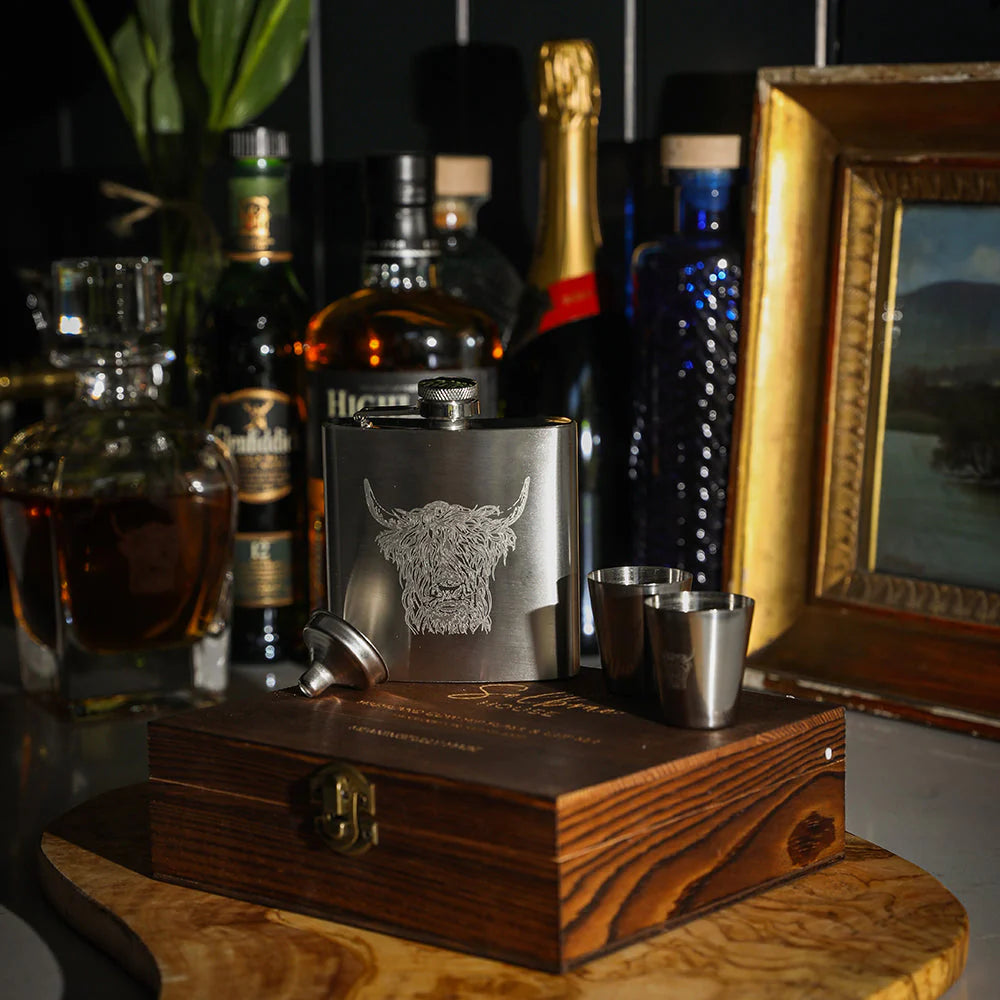A silver stainless steel flask engraved with a Highland cow head, presented on a wooden gift box with two matching metal shot glasses. The set is styled on a wooden table with whiskey and liquor bottles in the background.