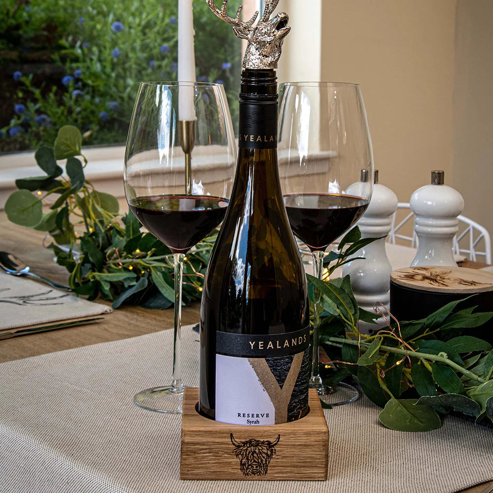Wine bottle with deer head stopper on a wooden stand, surrounded by glasses and decorative elements on a table.