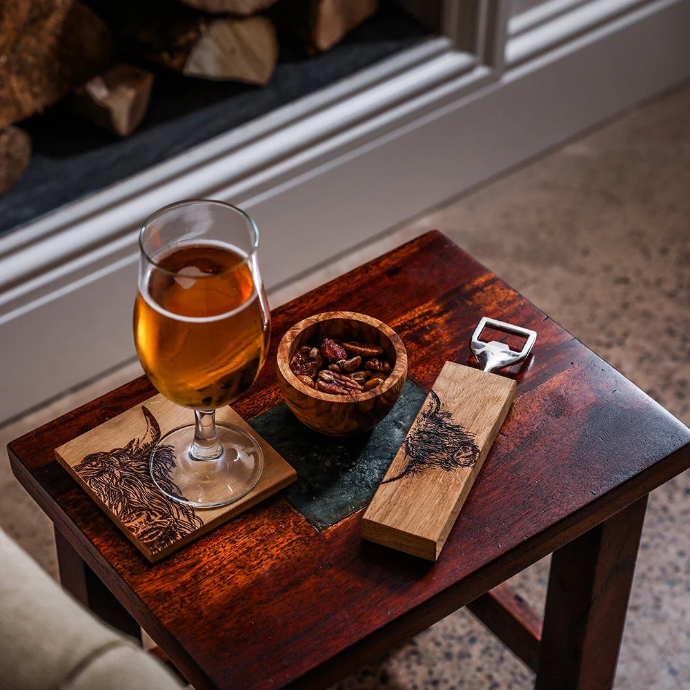 Glass of amber liquid on a wooden coaster with a bottle opener and small bowl on a wooden table.